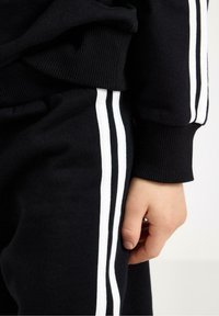 Černá mikina a kalhoty s žebrovanými manžetami, na bocích kalhot a rukávu tři bílé svislé pruhy. Ruka je viditelná.