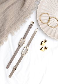 Deux montres avec un cadran rectangulaire ; l'une a un bracelet en silicone gris et l'autre en maille métallique. Des boucles d'oreilles en forme de cœur doré et une assiette beige à proximité.