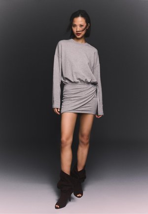 Femeie purtând o rochie gri, mulată, cu mâneci lungi și fustă drapată, și cizme negre, largi, cu toc înalt și degetele goale, stând pe un fundal întunecat.