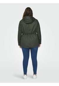 Dunkelgrüne Parkajacke mit tailliertem Bund, Kapuze und langen Ärmeln, kombiniert mit blauen, figurbetonten Jeans und weißen Turnschuhen. Glatte Textur.