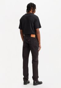 Hombre con cabello trenzado, vestido con camiseta negra, jeans oscuros con parche de cuero, y botas negras, de pie mirando hacia el lado en un fondo blanco.