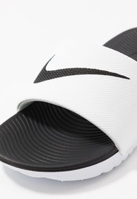 Mustat ja valkoiset liukusandaalit, joissa on teksturoitu kangasnauha ja uritettu kumipohja. Nauhassa on näkyvä musta swoosh-logo.