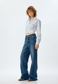 Camisa de rayas recortada en azul claro y blanco con mangas largas, combinada con unos pantalones vaqueros de pierna ancha y cintura alta en azul y unos zapatos marrones de punta.