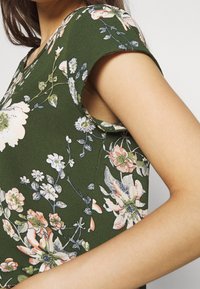 Jurk met bloemenpatroon op een groene achtergrond, korte opgerolde mouwen en een V-hals. Zachte stof met subtiele textuur en delicate bloemmotieven.