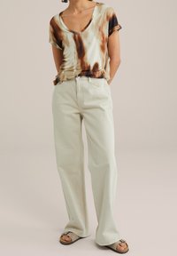 Beżowa bluzka z krótkim rękawem w stylu tie-dye z brązowymi wzorami, zestawiona z jasnoniebieskimi spodniami typu wide-leg; modelka w neutralnych sandałach.