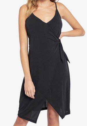 Kvinde iført en ærmeløs sort omslagskjole med sidetie og asymmetrisk kant, stående med den ene hånd på hoften.