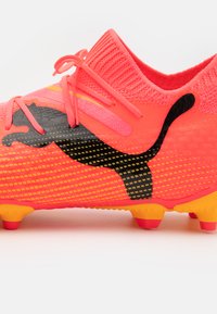 Scarpino da calcio di colore rosa brillante con parte superiore testurizzata, logo nero e accenti gialli. Presenta un colletto simile a una calza e tacchetti multidirezionali.