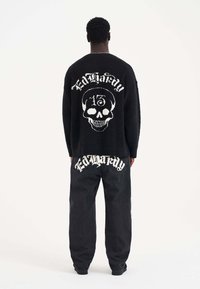 Zwarte fuzzy sweater met witte doodskop en "Ed Hardy" tekst op de achterkant, gecombineerd met losse zwarte jeans met "Ed Hardy" op de zakken.