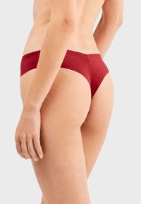 Tanga de encaje rojo con bordes de encaje, diseño sin costuras, tejido suave con acabado mate, corte bajo y mínima cobertura en la parte trasera.