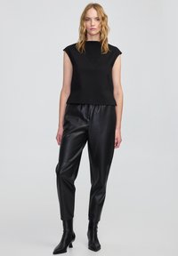 Schwarzes ärmelloses Oberteil mit strukturierter Passform und V-förmigem Ausschnitt, kombiniert mit hochtaillierten schwarzen Lederhosen und spitzen Stiefeletten.