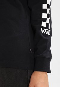Camiseta negra de manga larga hecha de algodón, con un patrón a cuadros blancos y el logo "VANS" en la manga derecha. Se puede ver una pequeña etiqueta del logo.