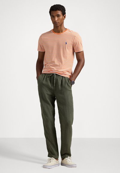 Πορτοκαλί και λευκή ριγέ κοντομάνικη μπλούζα σε συνδυασμό με πράσινα παντελόνια με κορδόνι. Η μπλούζα διαθέτει ένα μικρό μπλε λογότυπο; τα παπούτσια είναι ανοιχτόχρωμα.