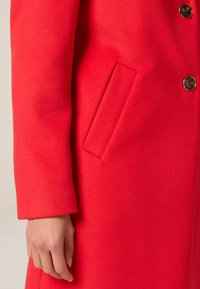 Abrigo de lana rojo con un acabado texturizado, que incluye un bolsillo lateral, cierre de botones y manga ligeramente ajustada.