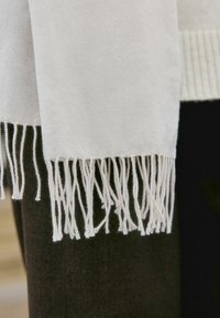 Hvid tørklæde med frynser i enderne, lavet af blødt materiale. Har en glat tekstur og et enkelt design, hængende over mørkt stof.