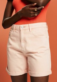 Shorts de suave denim rosa con puños enrollados, que presentan bolsillos frontales y un cierre de botón plateado. Top de textura acanalada en un brillante naranja.
