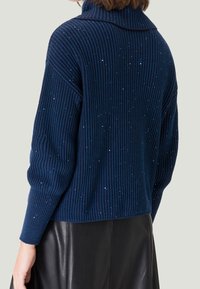 Mujer vestida con un suéter de canalé azul marino con pequeños brillos, combinado con una falda de cuero negra, vista por detrás.