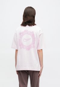 Άτομο με καστανά μαλλιά έως τους ώμους, φορώντας ένα φαρδύ ροζ μπλουζάκι με την κυκλική λογότυπο "Just do it" της Nike στην πλάτη.