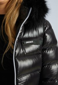 Blondýna v černé lesklé prošívané bundě s kapucí lemovanou kožešinou a bílým zipem, s nápisem "JACK1T" na hrudi.