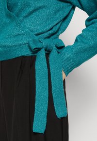 Teal stickad omslagströja med ett knytbälte, med en texturerad yta och ribbade ärmslut, bärs över svarta byxor.