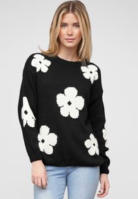 Un pull en tricot noir avec de grands motifs floraux blancs, un col rond et des manches longues, associé à un jean bleu clair.