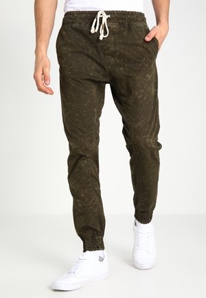 Uomo che indossa pantaloni jogger verde oliva con motivo, coulisse bianca e sneakers bianche, in piedi contro uno sfondo semplice.