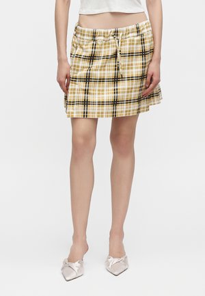 Femme portant une jupe à carreaux jaune et noire avec une taille élastique et des ballerines blanches pointues, debout devant un fond uni.