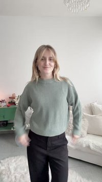 Mujer joven con cabello rubio, vestida con suéter verde y pantalones negros, de pie en una sala de estar luminosa con una decoración acogedora y una mesa auxiliar verde.