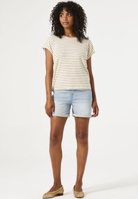 Chemise à rayures à manches courtes en crème et marron, accompagnée de shorts en denim bleu clair. Les accessoires comprennent des chaussures plates dorées et une posture décontractée.