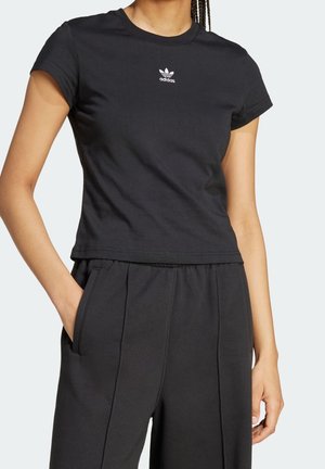 Person iført en sort kortærmet Adidas t-shirt og matchende sorte bukser med den ene hånd i lommen mod ensfarvet baggrund.