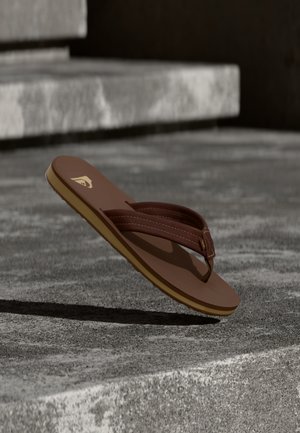 Pruun varbavõru sandaal kollase tallaga, hõljuv karedate betoonastmete kohal loomulikus valguses.