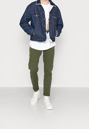 Giacca di jeans con colletto in shearling, T-shirt bianca, pantaloni verdi e sneakers bianche. Presenta tasche classiche e chiusura a bottoni. Vestibilità casual.