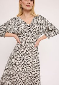 Czarna i biała sukienka w wzory z dekoltem w kształcie litery V, trzema ćwierć rękawami, guzikami z przodu oraz ściągniętą talią. Gładka tekstura materiału.