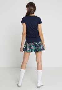 T-shirt blu navy abbinato a una gonna svasata con motivo floreale in tonalità scure, impreziosita da vivaci accenti tropicali. Calze bianche al ginocchio completano l'outfit.