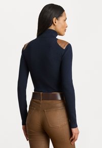 Tumman sininen ribbattu pooloneule, jossa on ruskeat nahkaolkapäät, yhdistetty ruskeisiin slim fit -haalareihin; tekstuuri parantaa visuaalista kontrastia.