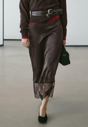 Brauner Satinrock mit Spitzenabschluss. Kombiniert mit einem dunkelbraunen Pullover und einem schwarzen Gürtel. Accessoires umfassen eine kleine schwarze Tasche und weiche schwarze Ballerinas.