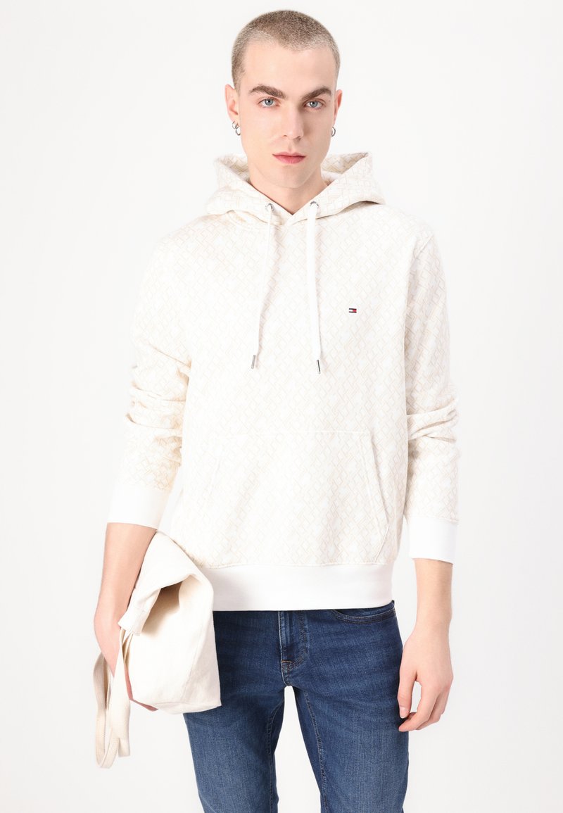 Cremefarbener Kapuzenpullover mit geometrischem Muster, vorderer Kängurutasche und weißen Bündchen; das Model hält eine hellfarbige Tragetasche.