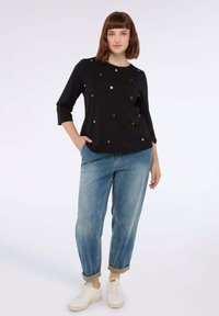 Maglietta nera a maniche lunghe con decorazioni metalliche, abbinata a jeans in denim azzurri arrotolati e sneakers bianche.