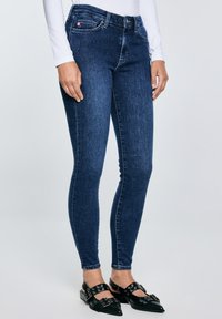 Žena u tamnoplavim uskim trapericama, bijeloj majici s dugim rukavima i crnim šiljastim cipelama s kopčom i detaljima od okruglica.