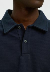Mornarska polo majica z strukturo ribane tkanine, klasičnim ovratnikom in dvema gumboma na vratu. Poglej bližje na podrobnosti majice.