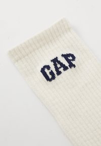 Hvit ribbestrikket sokk med marineblå "GAP"-logo strikket nær toppen av mansjetten på en enkel lys bakgrunn.