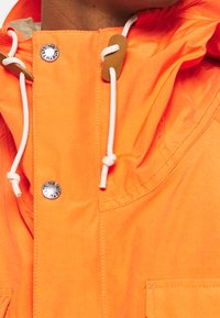 Oranžová nepromokavá bunda s kapucí, vybavená kontrastními bílými stahovacími šňůrkami a stříbrným cvokem. Viditelná texturovaná tkanina a detaily kapes.
