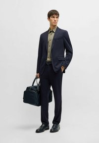 Marineblauer Anzug im Slim Fit, olivgrünes Hemd mit Knöpfen, schwarze Schuhe und marineblaue Tasche. Glattes Material, maßgeschneiderter Schnitt und minimalistisches Design.