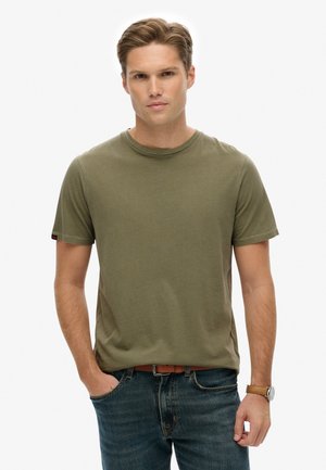 Ung mand med lyst brunt hår iført en olivengrøn kortærmet t-shirt, blå jeans, brun bælte og ur, stående med den ene hånd i lommen.