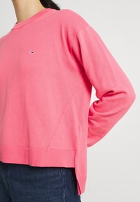 Rosa tröja med långa ärmar, ribbad nederkant, sidoflitsar och liten logotyp på bröstet; tillverkad av mjukt, lätt material.