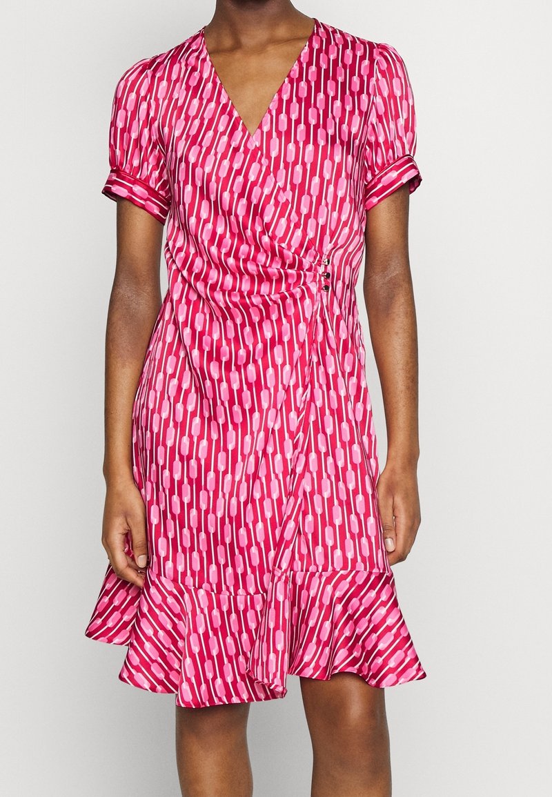 Femme portant une robe portefeuille rose mi-longue avec un motif géométrique, des manches courtes bouffantes et un ourlet volanté.