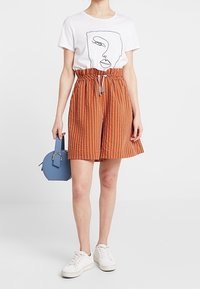 Camiseta blanca con diseño de arte lineal negro, combinada con pantalones cortos de cintura alta a rayas naranjas, zapatillas blancas y un pequeño bolso azul.
