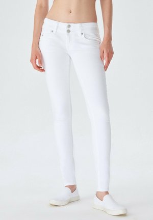 Kvinde model iført tætsiddende hvide jeans og hvide slip-on sko, står mod en ensfarvet hvid baggrund.