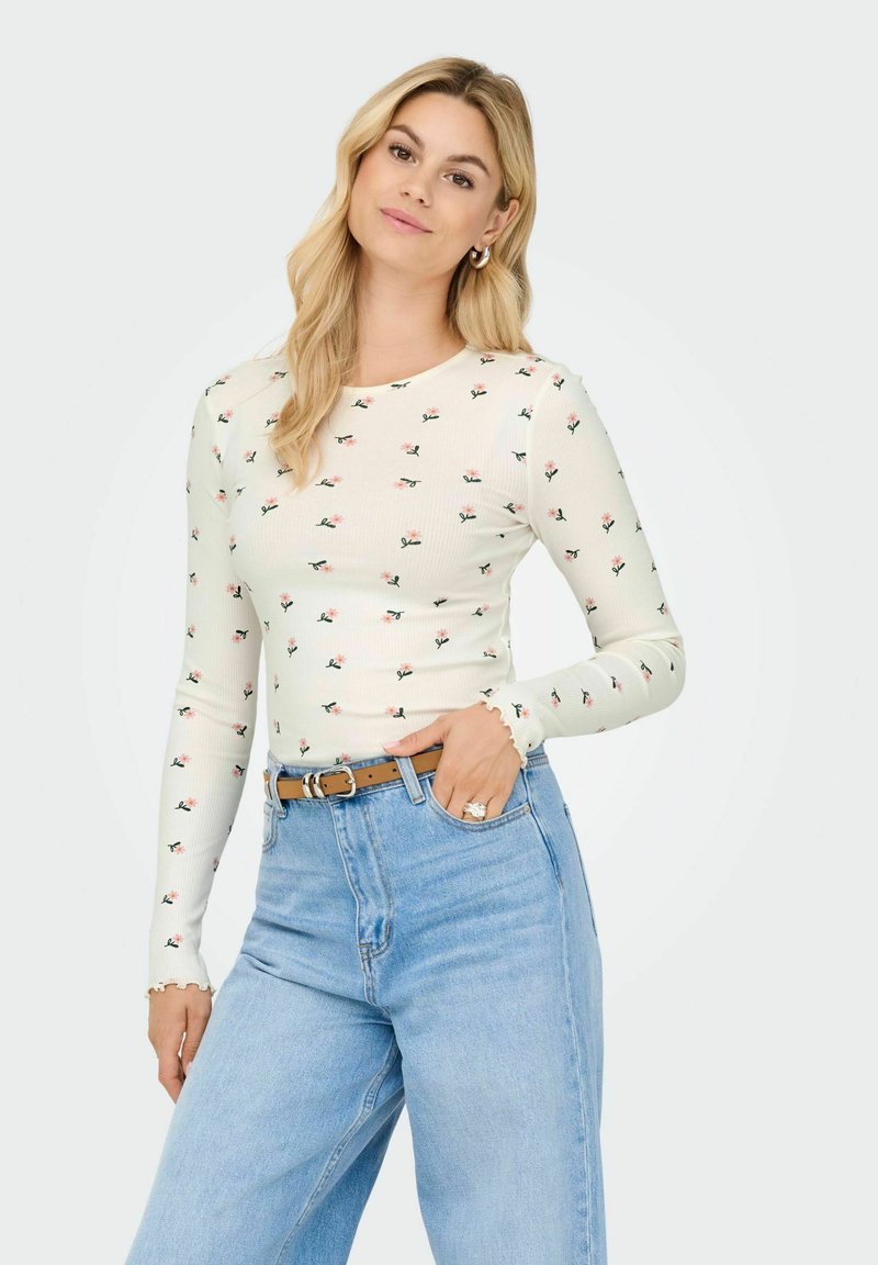 Hvid ribbet langærmet top med lille blomsterprint, rund halsudskæring og flæseærmer, parret med højtaffladede lyseblå jeans og et brunt bælte.