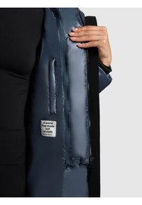 Tamsiai mėlyna pūkinė striukė su lygiu, blizgiu paviršiumi, su užtrauktuko kišene ir etikete, ant kurios parašyta „Šiltas apkabinimas iš audinio.“