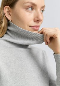 Grå ribbade polotröja med en åtsittande design, mjuk textur och hög krage. Modellen håller kragen med en hand.